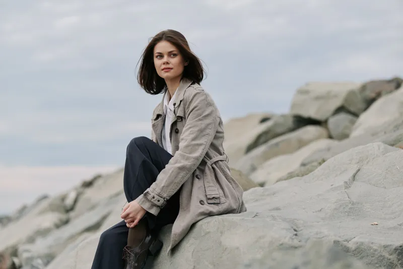 Young woman in casual outfit sits on large rocks by the sea, looking away thoughtfully. Overcast sky creates calm, reflective mood in natural outdoor environment.