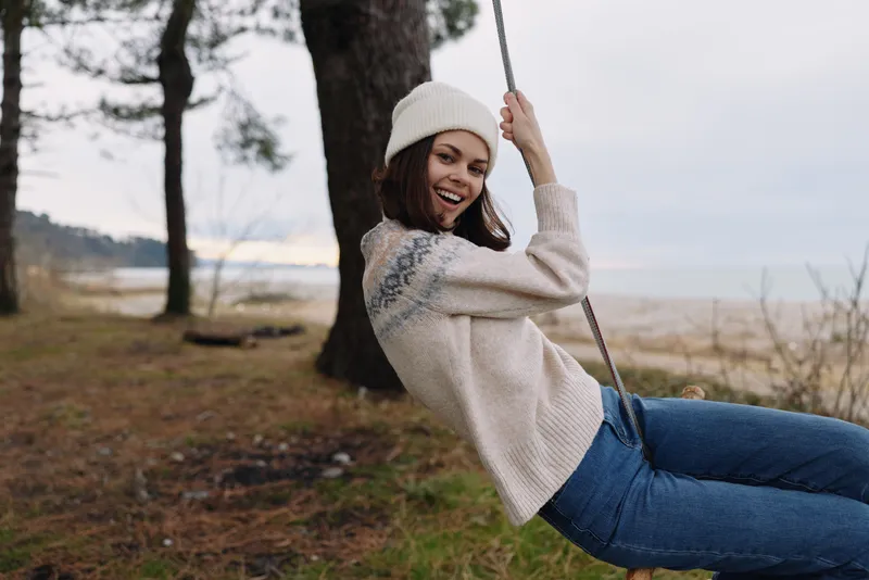 Young woman on a swing outdoors by a lake during daytime, wearing a knit sweater and beanie, smiling while enjoying a playful moment in a scenic park with trees and a calm shoreline.