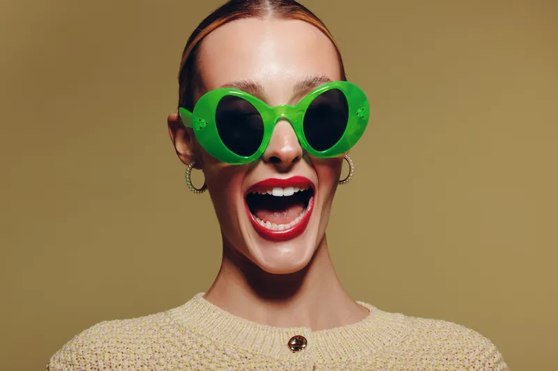 Happy woman with bright red lipstick wearing large green sunglasses and beige sweater smiles widely on neutral background. Concept of fashion, joy, and vibrant style portrait.