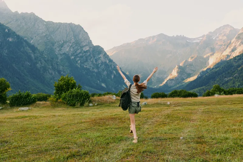 Joyful woman outdoors in a field near mountains, arms raised, backpack ready, sunlit moment captures freedom, exploration, and a casual travel lifestyle