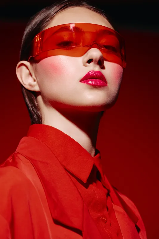 Young woman in red futuristic eyewear and red fashion shirt poses confidently against a red background. Creative beauty portrait with bold makeup concept.