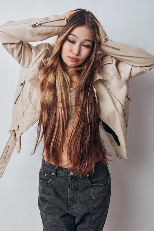 Young woman with long hair wearing jacket and dark jeans posing against white background with eyes closed and hands on head in casual fashion portrait indoor.