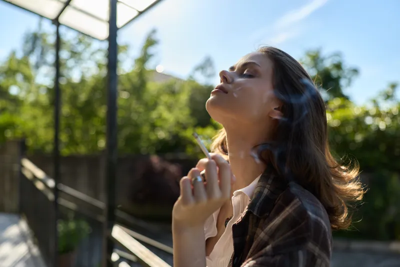 Young woman tilts her head back in sunlight, eyes closed, savoring a warm outdoor moment in a plaid shirt with a relaxed, peaceful vibe, greenery and clear sky behind her