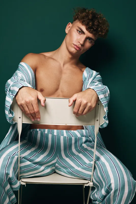 Young man with curly hair wearing striped pajamas sitting backwards on chair against dark green background. Casual pose, relaxed mood and fashion style with bare chest and open shirt.