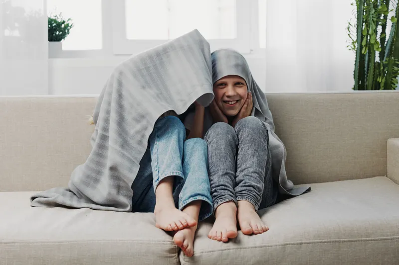 Two happy children sitting on sofa covered with blanket smiling and having fun in bright cozy living room at home.