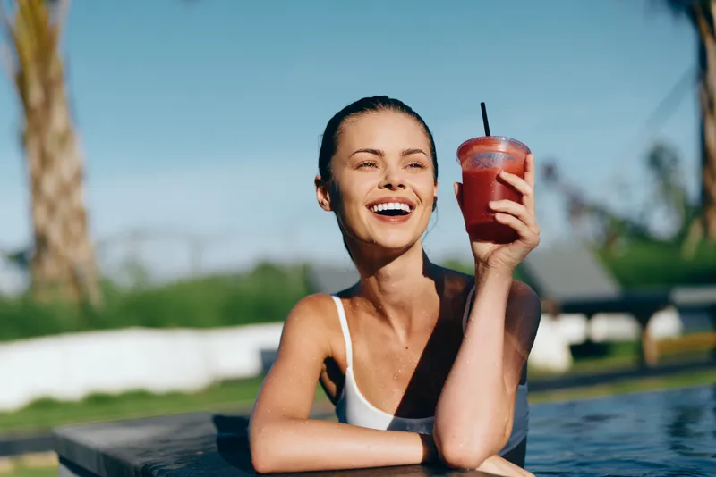 A smiling woman lounges by a pool with a bright red drink, enjoying a refreshing moment in warm sun. Concept: relaxation and carefree summer lifestyle.