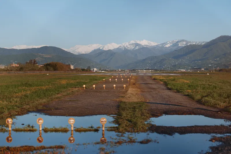 Dirt path leads through grassy fields toward distant mountains with snow capped peaks and reflective puddles under a clear blue sky, creating a tranquil outdoors moment.