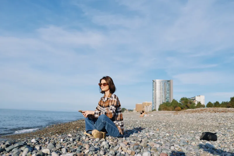 Beach scene with a woman sitting on pebbles by the ocean, wearing a plaid shirt and jeans, sunglasses, in a relaxed pose, distant urban buildings under a clear sky