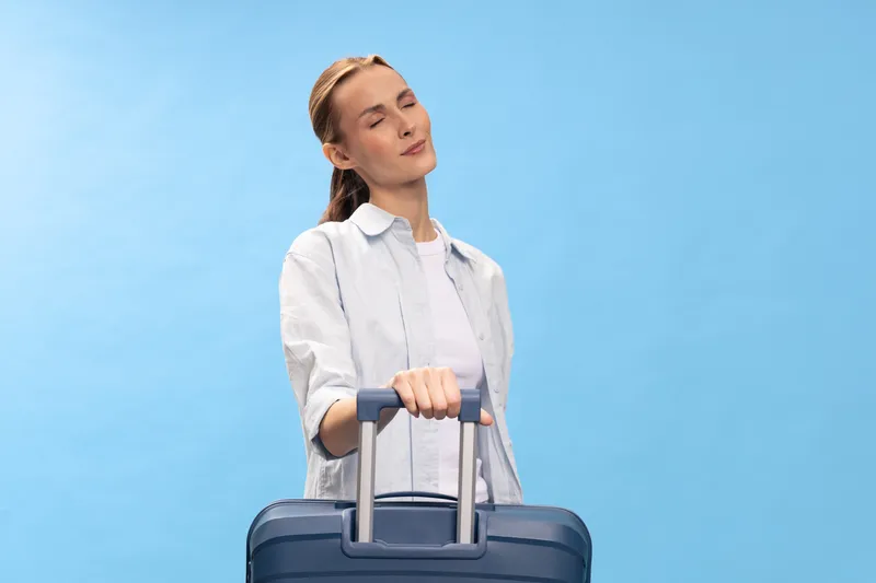Young woman with closed eyes holds suitcase handle, standing against blue background. Concept of travel, vacation, relaxation, and leisure time for vacation planning.