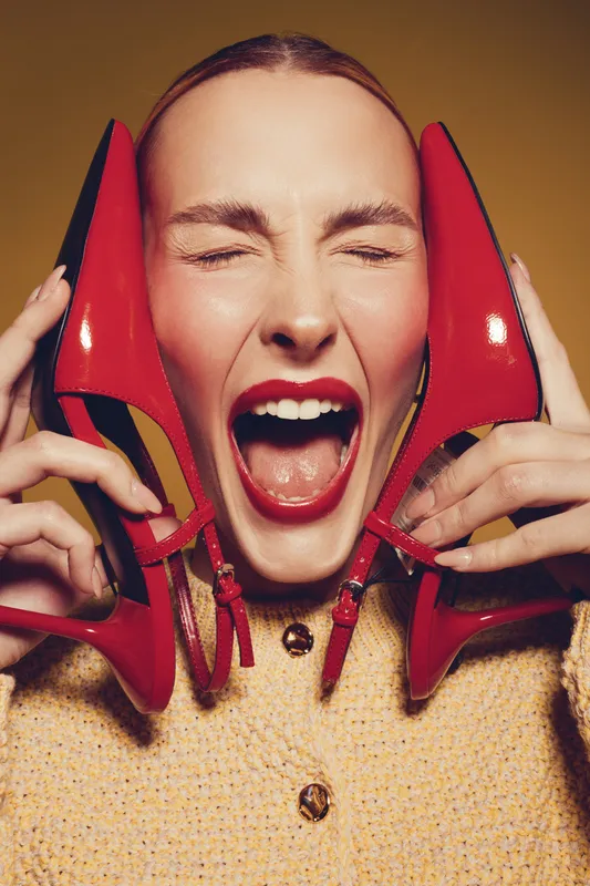 Young woman with closed eyes holds red high heels near face, mouth open in excitement. Fashion concept with bright makeup and joyful expression on beige background.