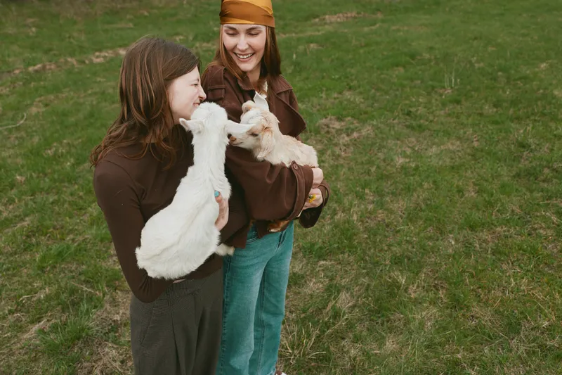 Two young women smile while holding baby goats outdoors on a grassy field during a relaxed lifestyle moment in casual clothing on a cool day.