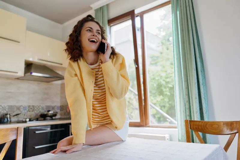 A woman in a yellow cardigan chats on the phone in a kitchen, smiling, conveying a casual, happy home lifestyle indoors, with warm interior details and daylight through a window.