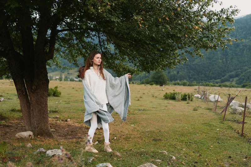A woman stands in open countryside, wearing a light shawl and casual jeans by a large tree, conveying a calm, contemplative moment in a rural lifestyle setting.