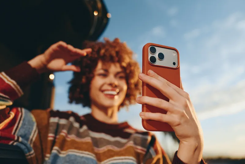 Woman takes selfie with smartphone outdoors, smiling with curly hair, holding phone close to camera, casual striped sweater, warm sunset light, sky background, lifestyle moment