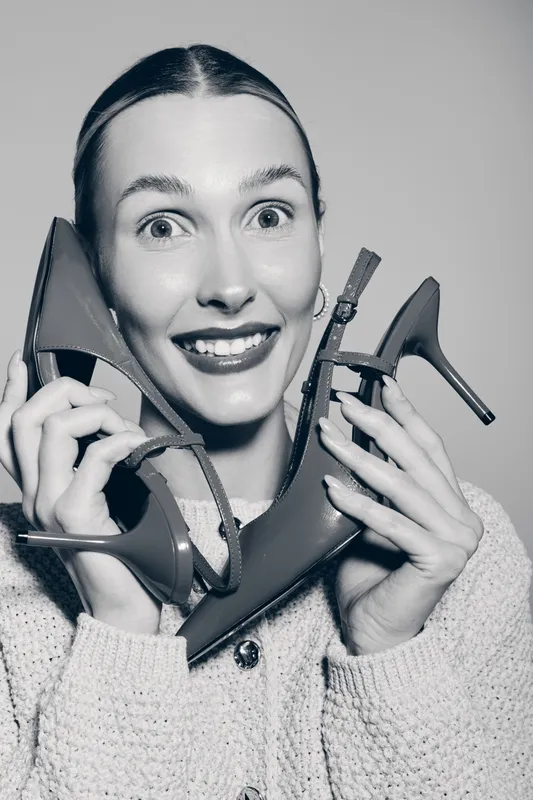 Young white woman with pale skin smiling and holding pair of high heels near face wearing knitted sweater.