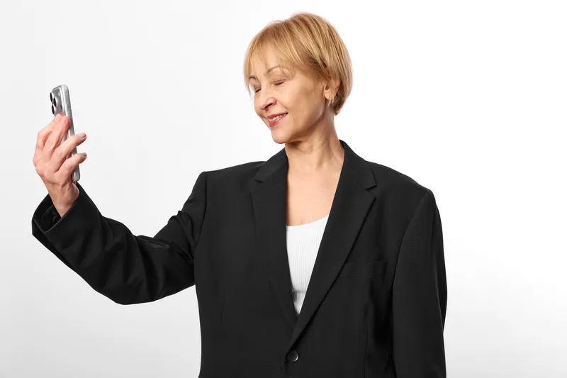 Senior woman holding a smartphone, short blonde hair, slight smile, looking at the screen, white studio background, technology and communication, soft light