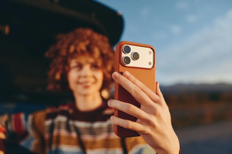 Woman holding smartphone for a selfie, hand close up with phone in outdoors lifestyle scene. Blurred curly hair woman in background on street at sunset, soft natural light.
