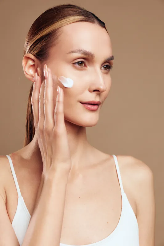 Young woman applies moisturizing cream on face with fingers looks focused skin care beauty routine isolated on beige background natural healthy complexion closeup portrait
