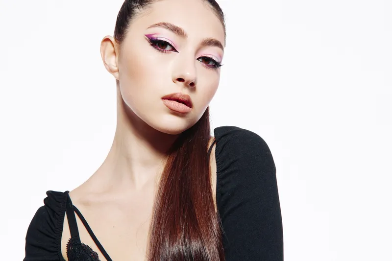 Young woman with sleek ponytail and bold pink eyeliner wearing a black top poses against white background. Concept emphasizing beauty, fashion, and creative makeup.