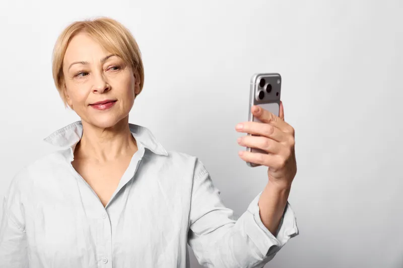 Senior woman holding a smartphone for a selfie, calm expression, indoor neutral background, one hand raised, lifestyle concept of digital communication today