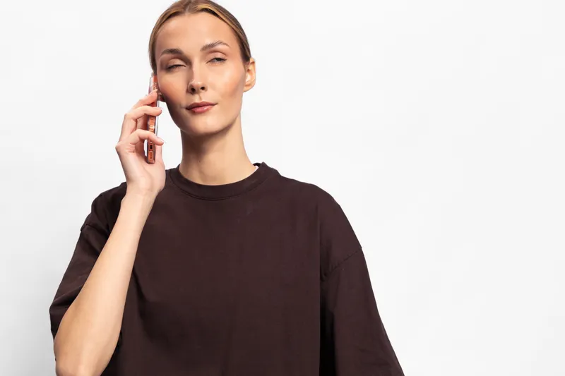 Young woman in dark loose shirt holds smartphone near ear with closed eyes. White background and soft lighting create calm atmosphere. Concept of conversation, communication, and concentration.