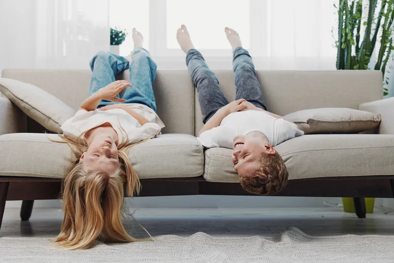 Two children lying upside down on a beige sofa in casual clothes, smiling and enjoying playful moment in a bright living room with natural light and green plants.