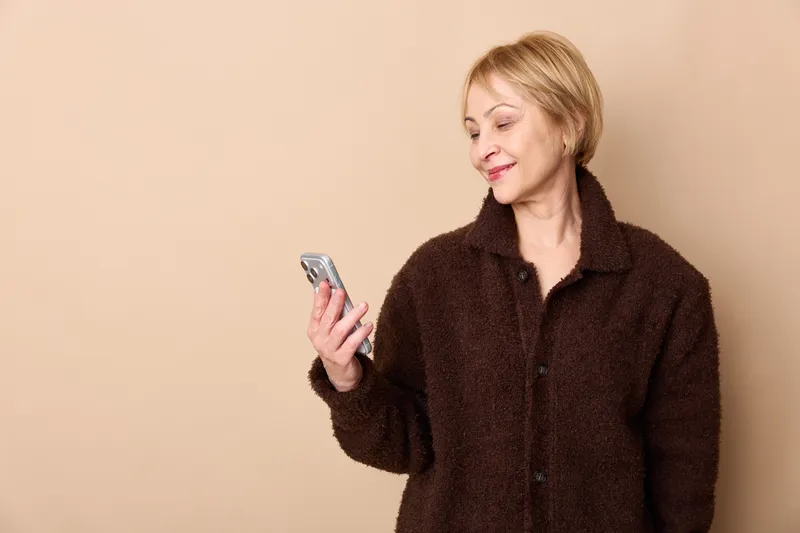 Elderly woman smiling indoors holding smartphone in relaxed portrait pose on beige background soft warm light casual sweater calm expression with gentle gaze for mobile use
