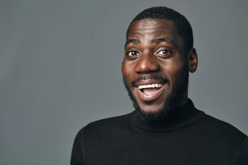 African man with dark skin smiling and looking at camera in creative conceptual visual style. Portrait of happy male with beard wearing black turtleneck on gray background.