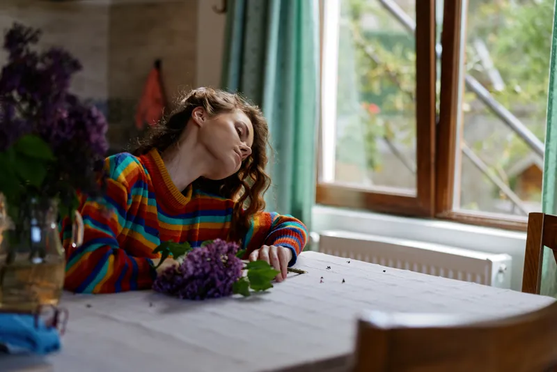 Woman relaxing indoors at a cozy home table, soft natural light from a window, wearing a colorful sweater with fresh flowers, capturing a calm lifestyle moment with a peaceful mood and warm colors