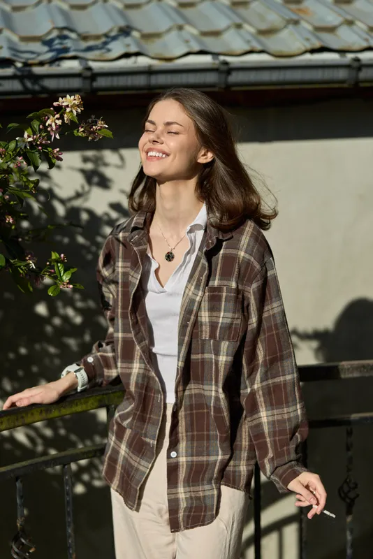 A relaxed woman in a plaid shirt stands outdoors, sunlit and smiling, wearing a white tee and beige pants. Lifestyle portrait capturing casual confidence and warmth