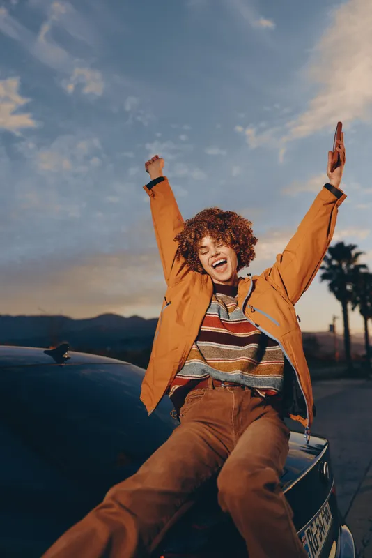 Woman curly hair laughing, jacket, car, sunset, lifestyle, outdoors, travel, hands raised, joyful portrait, tan outerwear, soft evening light