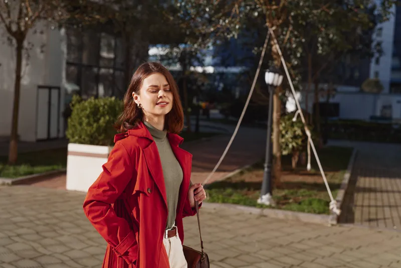 A stylish woman in a red coat stands outdoors on a sunny day, eyes closed against the glare, embodying urban fashion, confidence, and seasonal style.