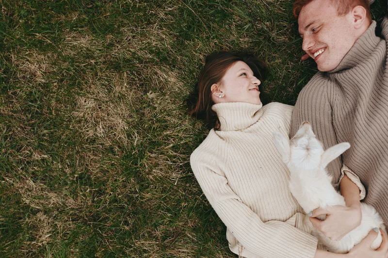 Joyful couple outdoors sharing a tender moment on grass with a small goat kid, cozy sweaters, soft light, relaxed mood, candid scene, warm tones, gentle breeze, natural setting romantic mood,
