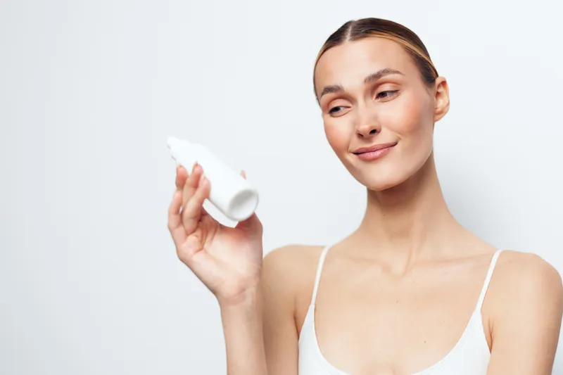 Woman holding a white cup for skincare beauty in a studio portrait, gentle smile, clean minimal background, young face, white tank top, soft daylight, copy space