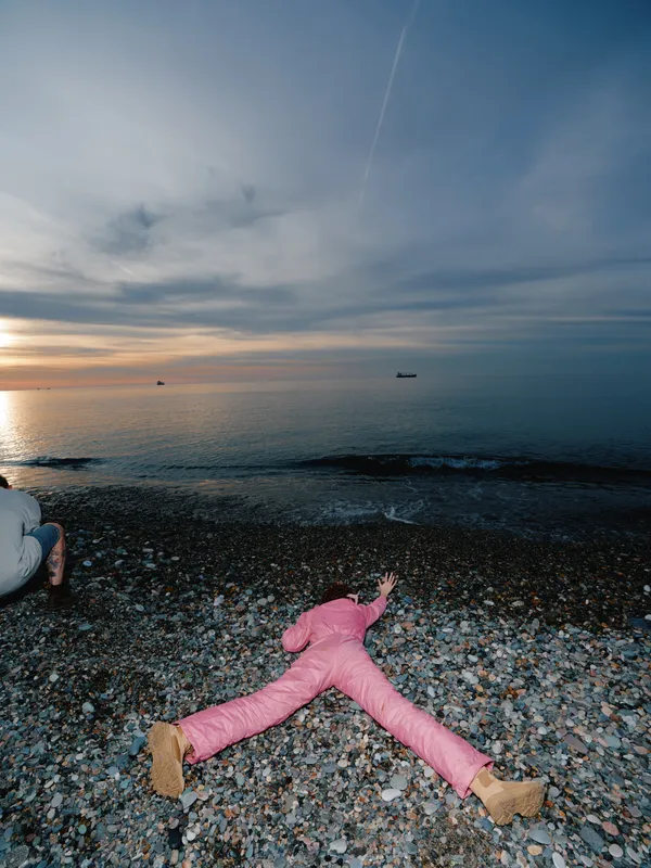 Beach scene with a person lying on a pebbled shore at sunset, arms and legs spread, calm sea and distant boats, pink outfit, dramatic sky, tranquil mood.