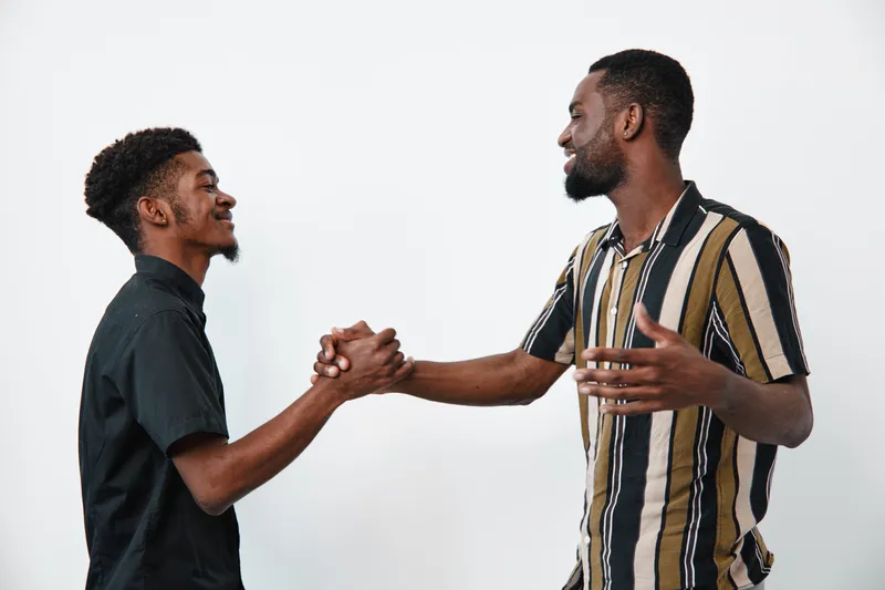 Two black men with dark skin shaking hands and smiling in casual clothes on white background. African ethnicity, friendship, greeting, connection, communication and positive interaction.