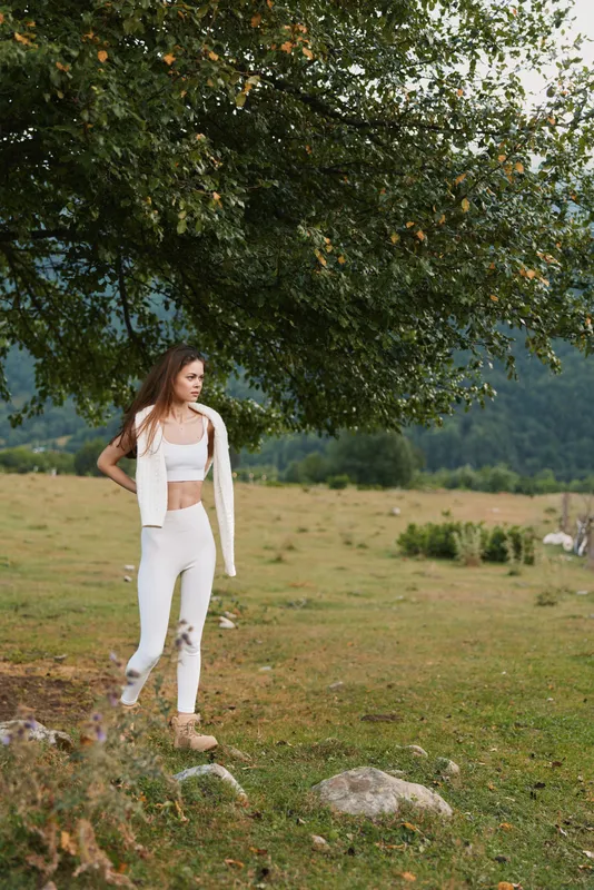 Young woman in white croptop and cardigan stands in a sunlit meadow beside a tree, calm stride, casual style, peaceful countryside mood
