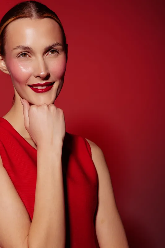 Smiling woman with red lipstick and red sleeveless top poses with hand near chin against red background. Concept of beauty, confidence, fashion, and portrait photography.