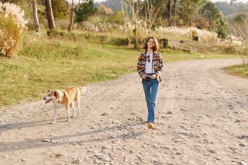 Woman walking with a dog along a gravel path in a sunlit countryside, casual outfit, open fields and a relaxed mood, capturing a peaceful outdoor moment on a clear day.