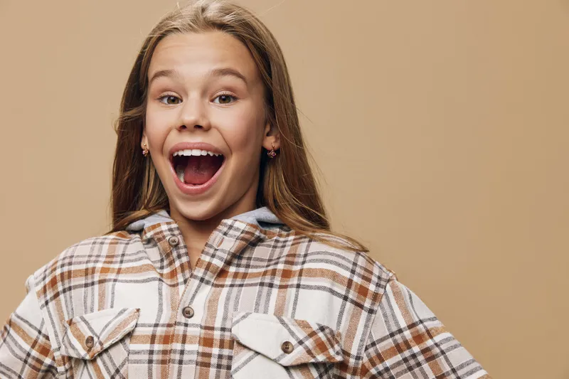 Happy teenage girl smiling with open mouth wearing plaid shirt on beige background. Excited female teenager with long hair, casual fashion and joyful expression in studio portrait.