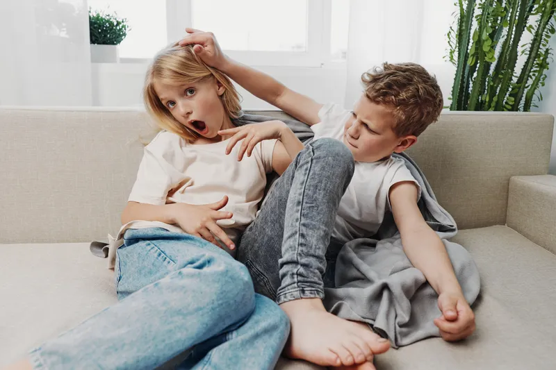 Two children in casual clothes are playfully fighting on a couch in a bright living room. The boy and girl show surprised and focused expressions during their interaction.