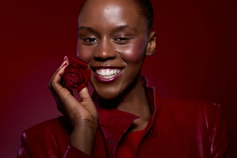 Smiling African woman with dark skin holds red rose near face, wearing red leather jacket on red background. Concept of beauty, happiness, romance, and fashion with vibrant colors.