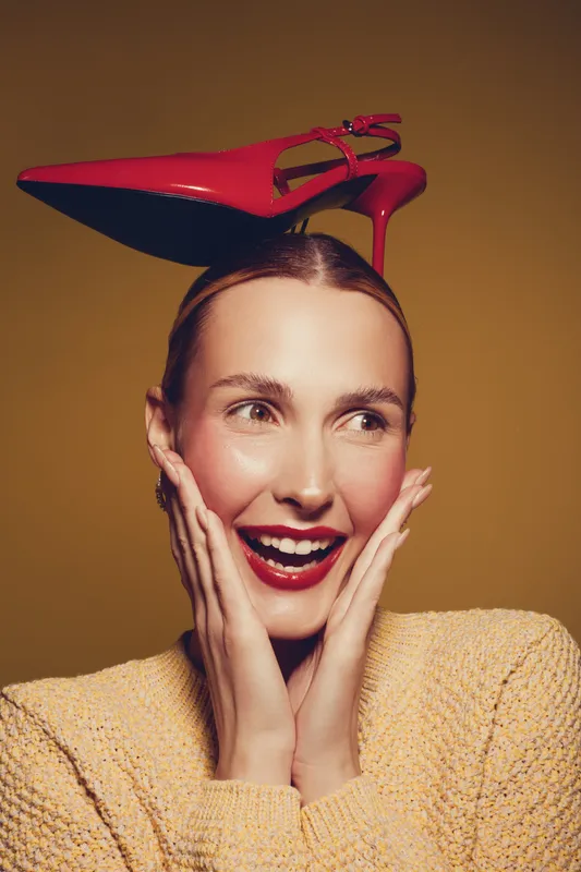 Smiling woman with red high heel shoe on head looks surprised and happy. She wears a yellow sweater and bright lipstick on brown background. Concept of fashion and fun.