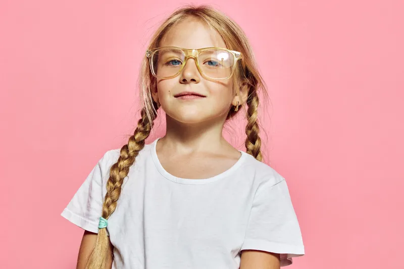 Smiling young girl with glasses and braided hair wearing white t shirt on pink background.