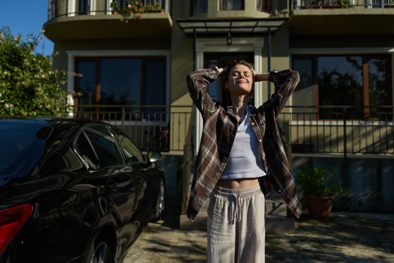 Young woman outdoors, arms raised in a sunlit moment, wearing a plaid shirt and gray joggers, radiating relaxed confidence and candid happiness in a casual lifestyle scene