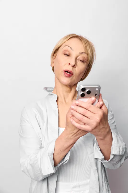 Elderly blonde woman holding smartphone, surprised expression, looking at screen, casual white shirt, isolated on white background, vertical studio portrait
