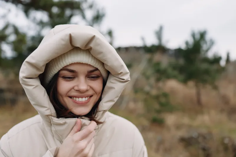 Woman wearing a beige hooded jacket outdoors, smiling softly in a natural field with sparse trees, capturing a calm, cozy moment in autumn air and gentle light.