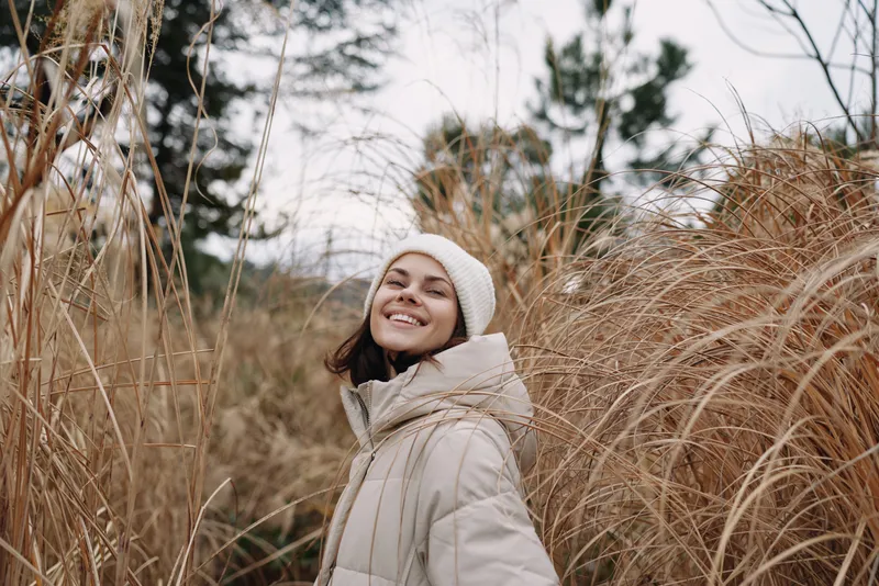 Woman outdoor winter coat smile nature among tall dry grasses in a natural landscape, capturing a warm candid portrait in a serene meadow scene.