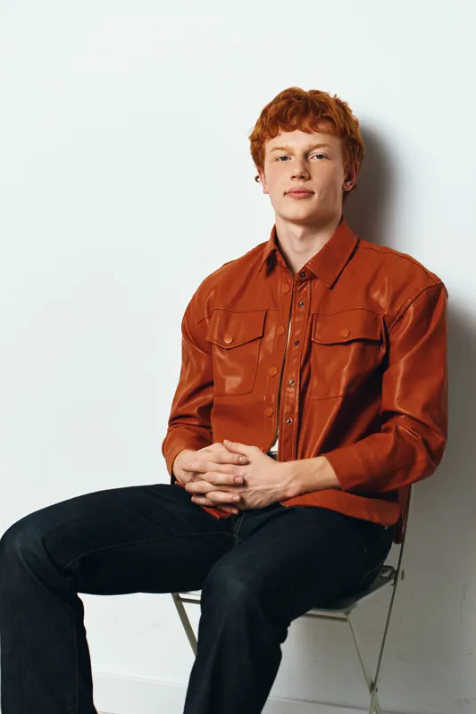 Young man with red hair sitting on chair against white background wearing orange shirt and black pants with hands clasped in front of him casual posture and direct gaze.