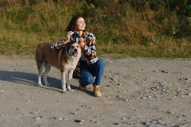 Woman outdoors with a dog on a gravel beach, crouching and smiling while holding a camera, capturing candid moments in sunny natural light.